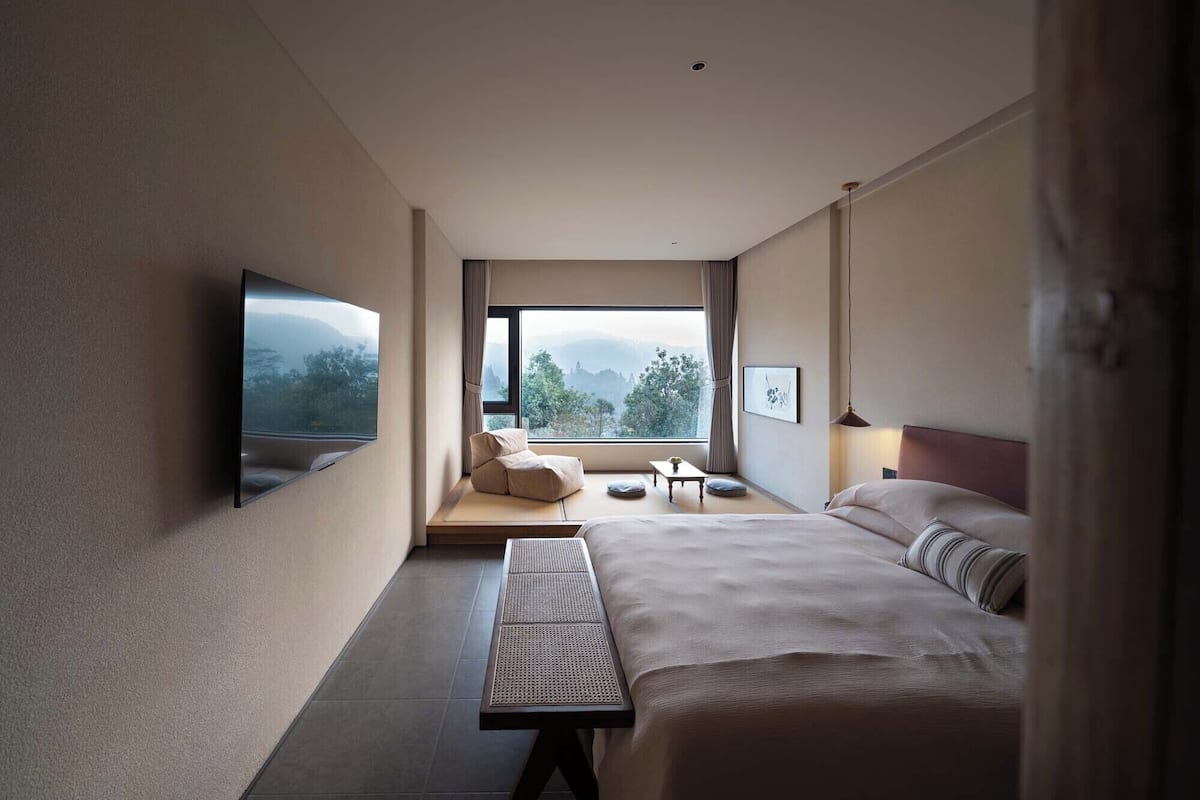 Una habitación de hotel moderna con una cama grande, un televisor de pantalla plana en la pared, una mesita con una silla y un ventanal con vistas al exterior.