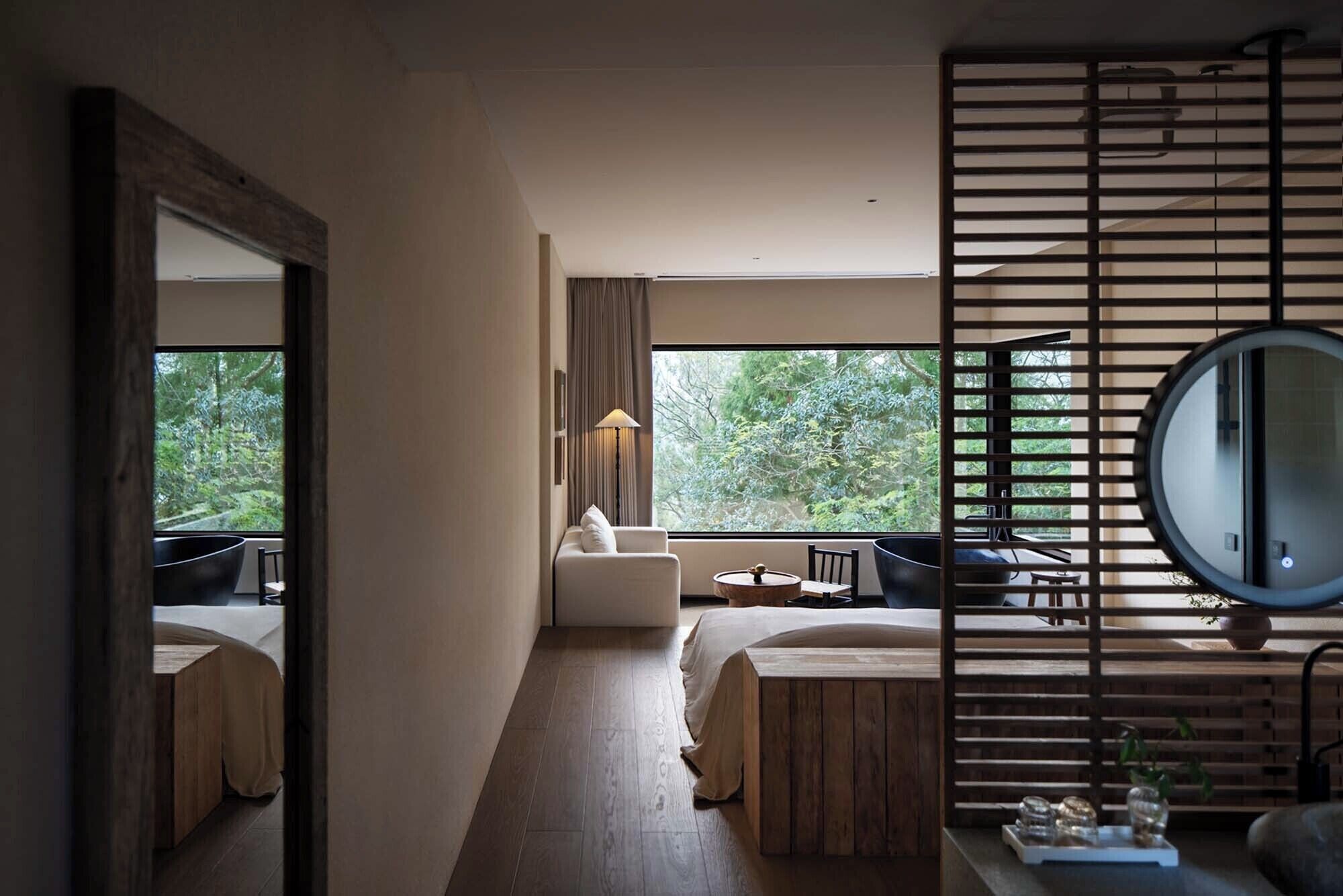 Une chambre d’hôtel moderne avec un grand lit, un petit canapé, un miroir rond et une vue sur l’extérieur.