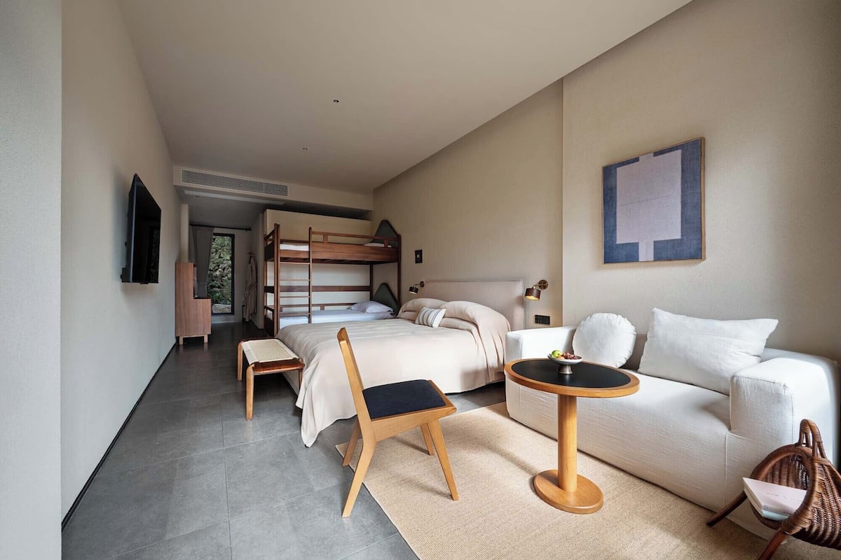 Una habitación de hotel moderna con una cama litera, un sofá, una mesa de centro redonda, una silla y un televisor.