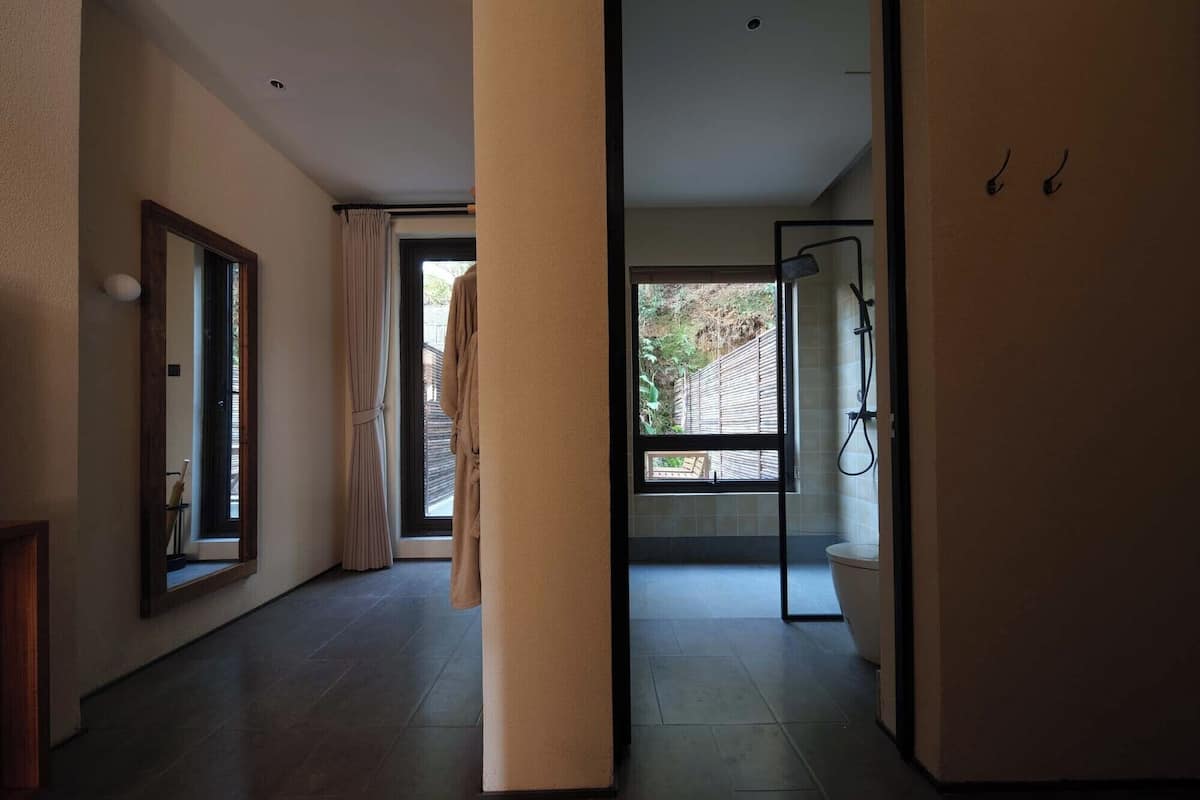 Un baño moderno con cabina de ducha de vidrio, inodoro y ventana con vista a una terraza de madera y follaje.