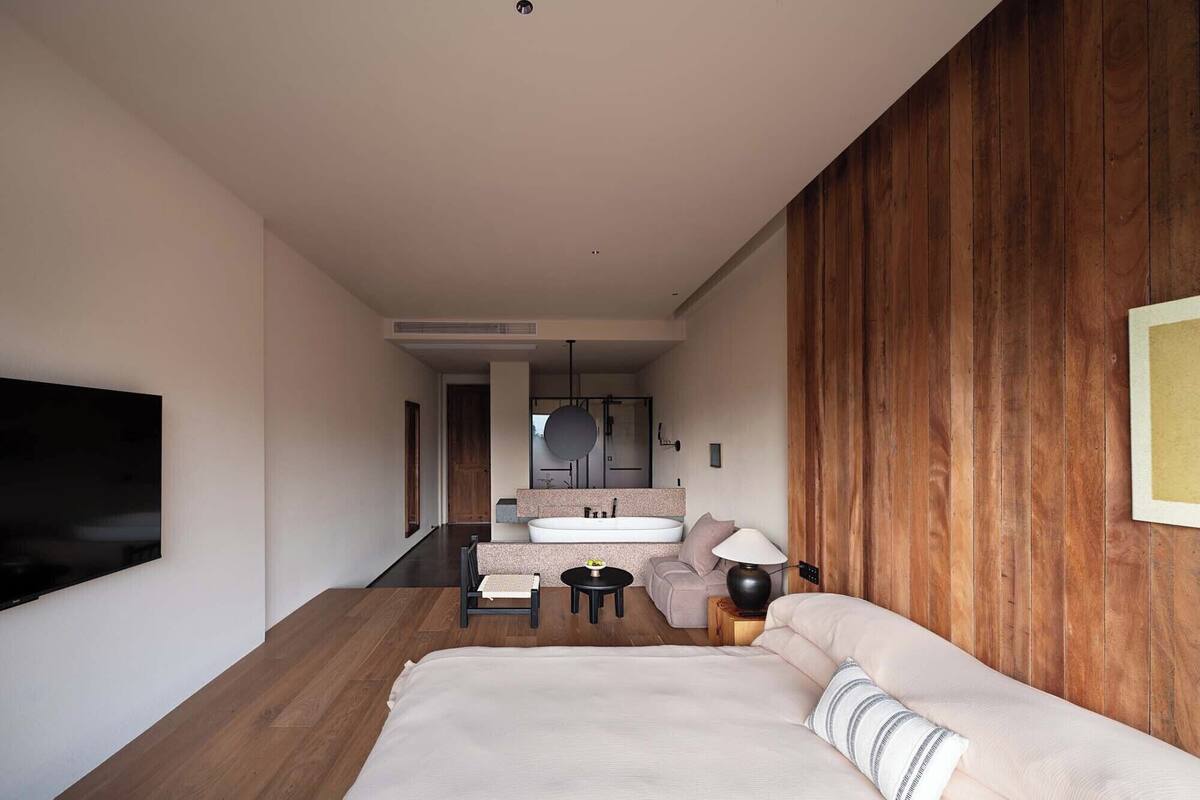 Un dormitorio moderno con una cama grande, una pared de acento de madera, un televisor montado en la pared y un baño visible a través de una puerta abierta.