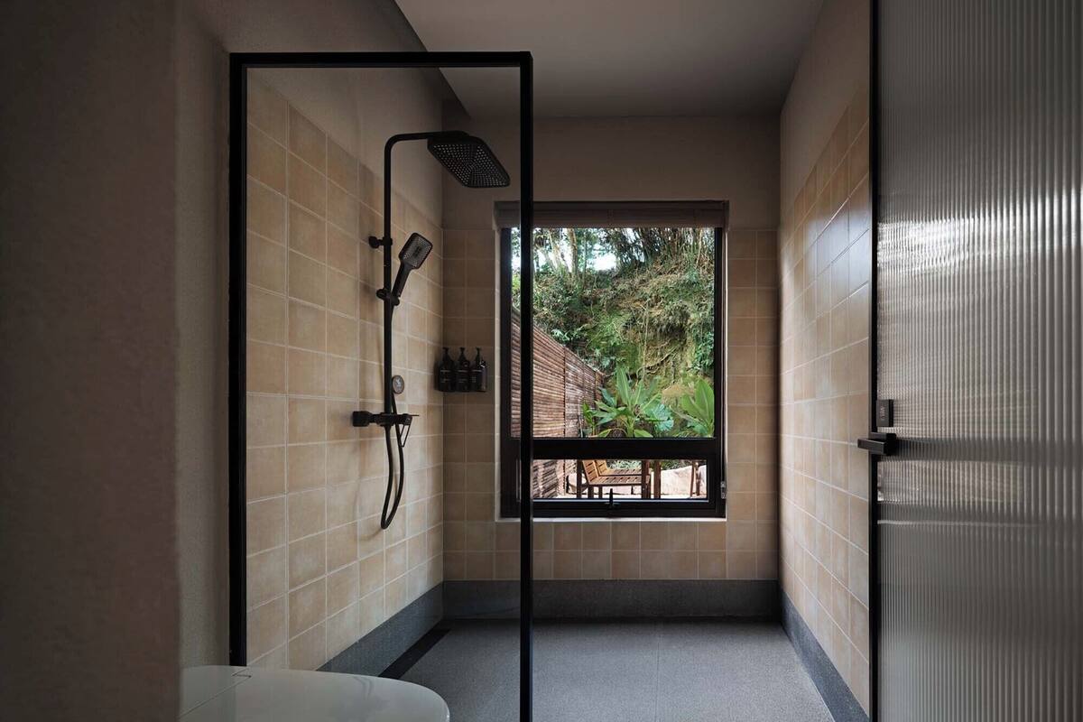 Un baño moderno con cabina de ducha de vidrio, vista a un jardín a través de una ventana y un lavamanos blanco.