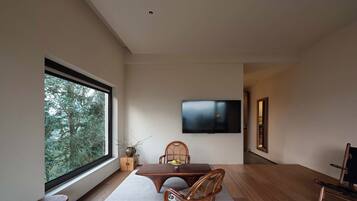 Una sala de estar moderna con un ventanal grande, un televisor de pantalla plana montado en la pared, una mesa de comedor con sillas y un taburete pequeño.
