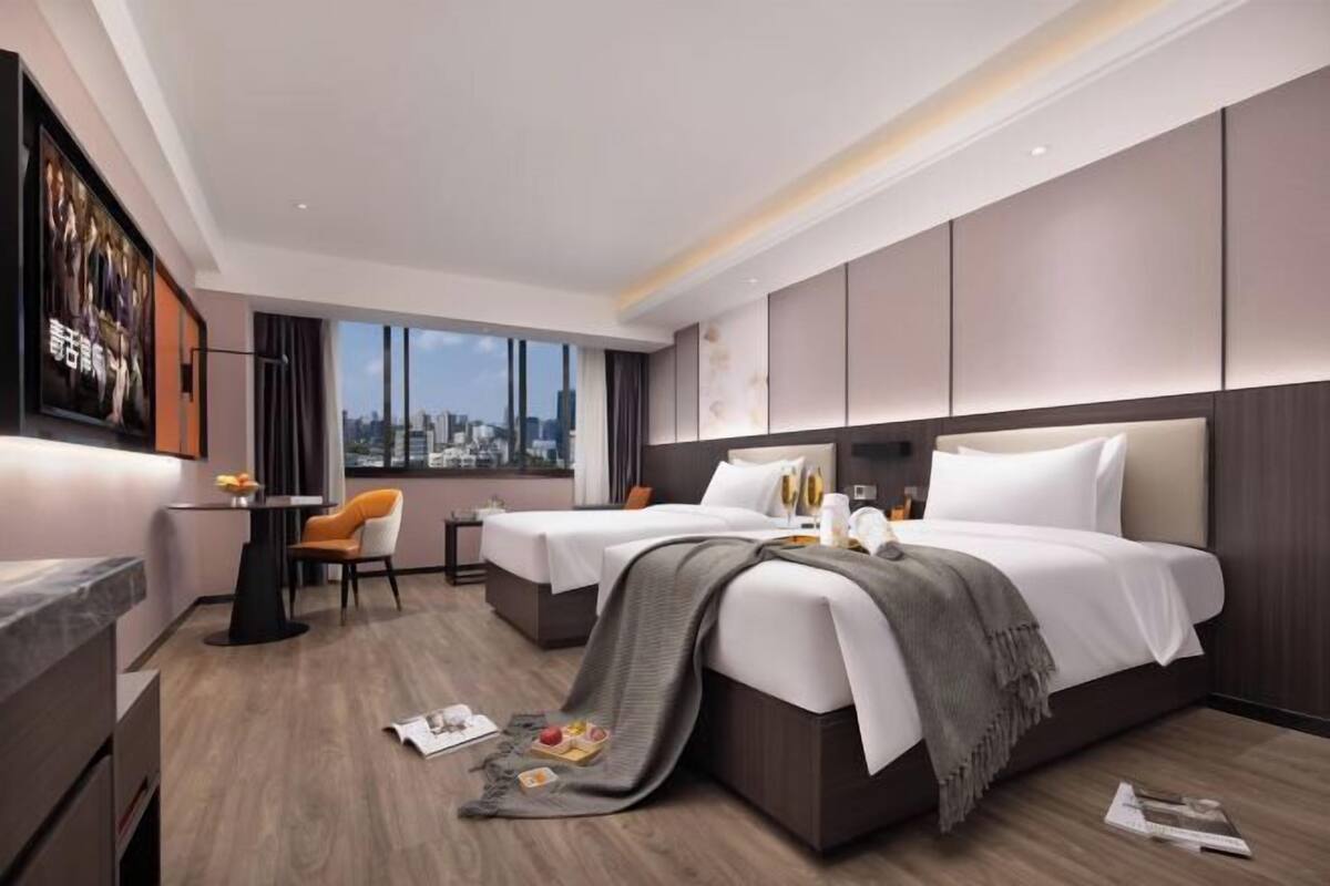 A modern hotel room with two beds, a dining area, a large window with a city view, and a mounted television.