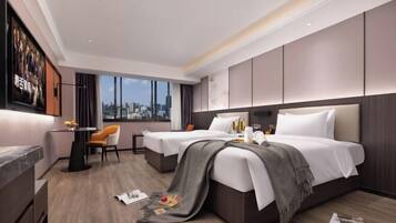 A modern hotel room with two beds, a dining area, a large window with a city view, and a mounted television.