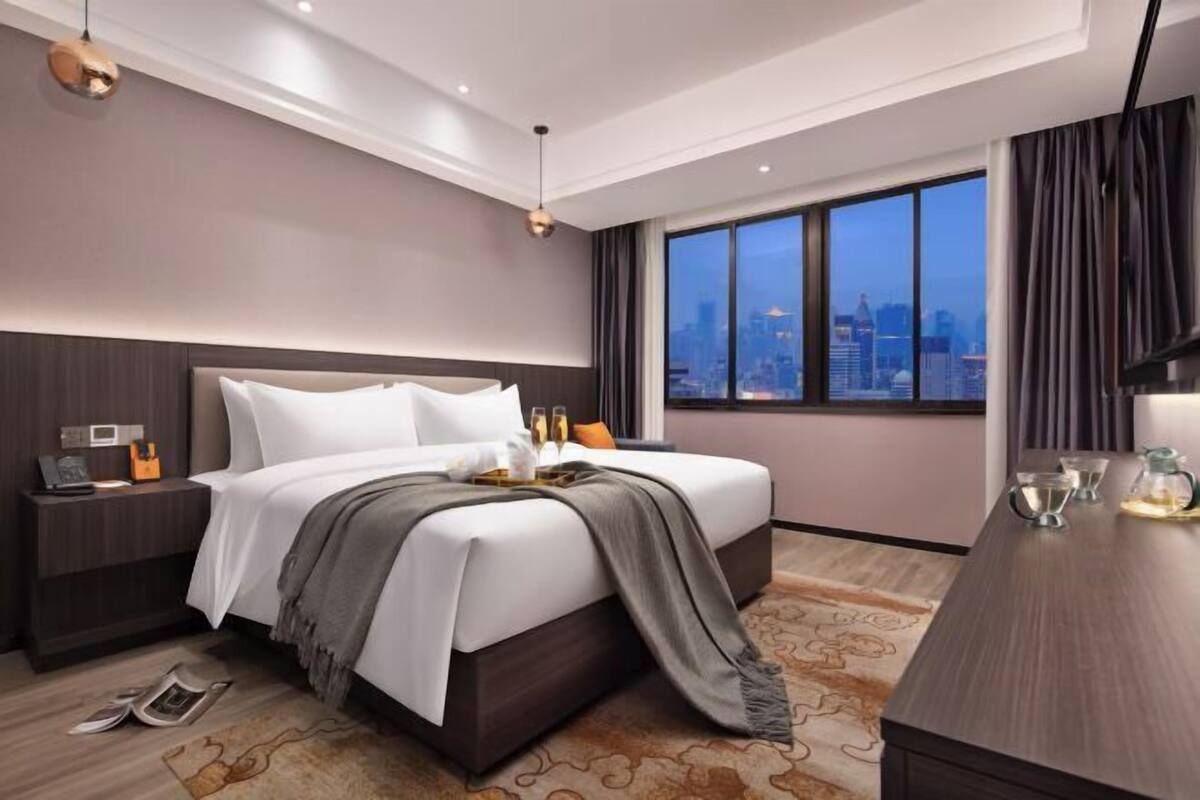 A modern hotel room with a large bed, a nightstand, a desk, and a city view.