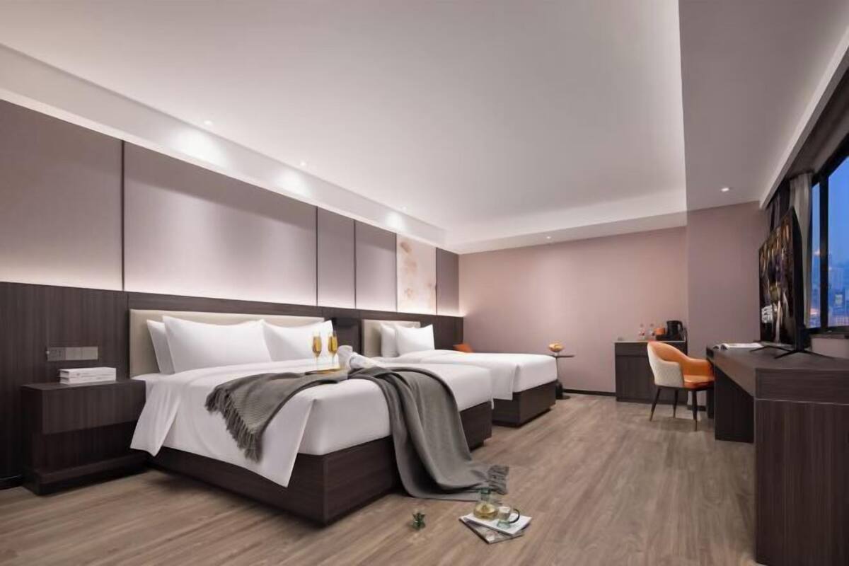 A modern hotel room with two beds, a desk, and a view of the city.