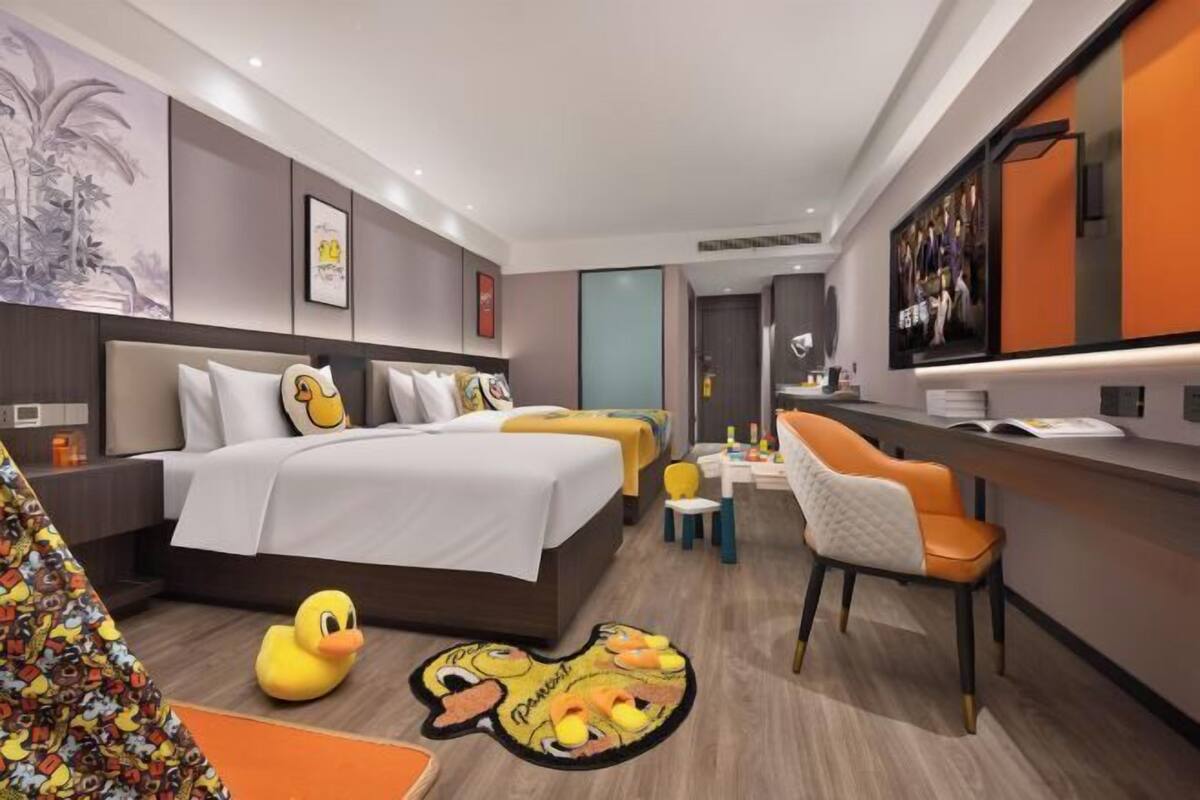 A modern hotel room with a large bed, a desk, and a chair. There are decorative pillows and a duck-themed rug.