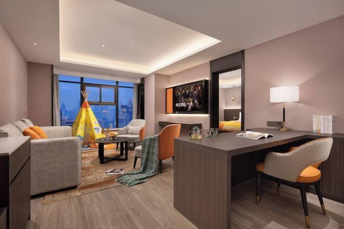 A modern hotel room with a large window, a desk, two chairs, a sofa, and a decorative teepee.