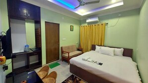 A hotel room with a bed, a desk, a chair, a ceiling fan, and a window with curtains. - Hotel Aayes & Resort (Bolpur)