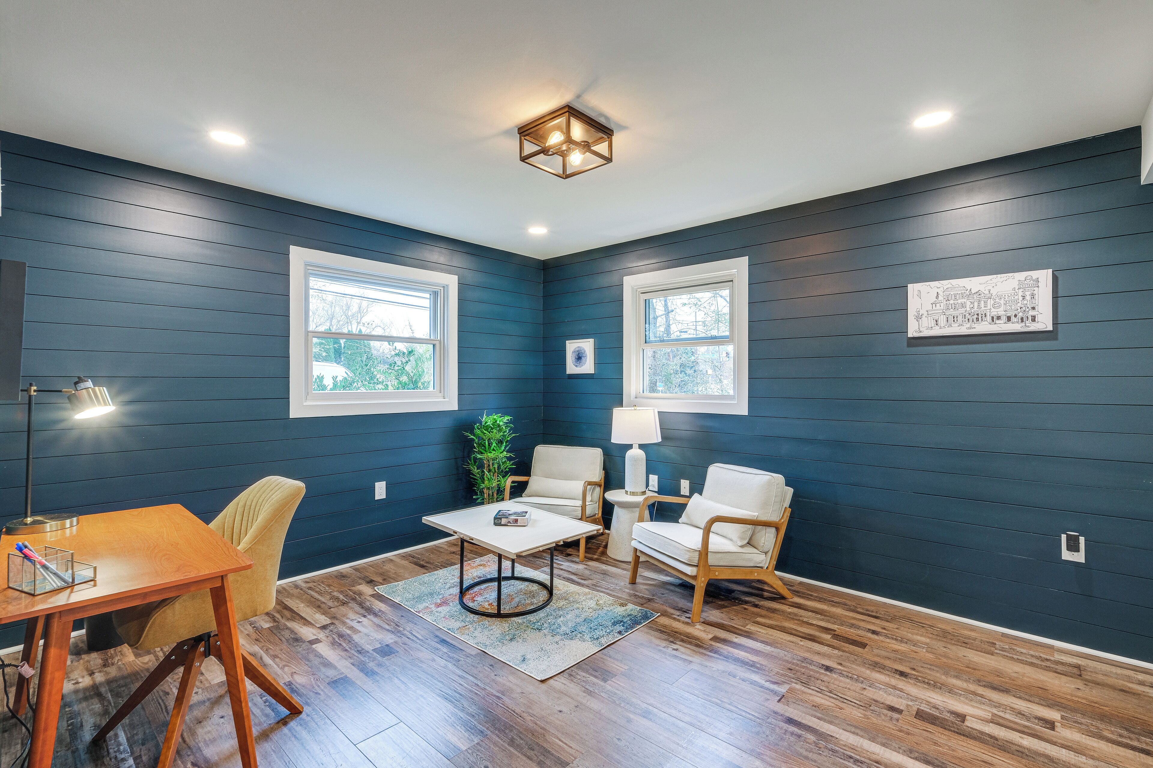 Une pièce aux murs bleu foncé, dotée d’un plancher en bois, d’une table à manger avec des chaises, d’une table basse, d’une lampe et d’un tableau encadré accroché au mur.