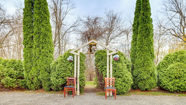 Un vialetto conduce a un arco di legno con due sedie ai lati, circondato da siepi curate e alberi alti.