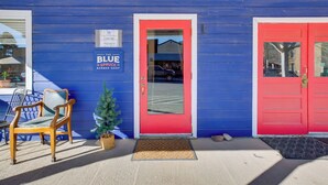 Un edificio azul con una puerta roja y una puerta azul. Hay una silla y un pequeño árbol de Navidad afuera.
