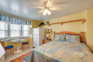 Une chambre à coucher comprenant un lit, un ventilateur de plafond, une fenêtre avec des stores, une petite table avec des chaises, une table de chevet et une étagère avec des objets décoratifs.