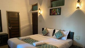 A double bed with a white and green floral bedspread, two pillows, a bedside table with a phone, and a wall-mounted light fixture. - Indraprasth Heritage Resort (Gogunda)