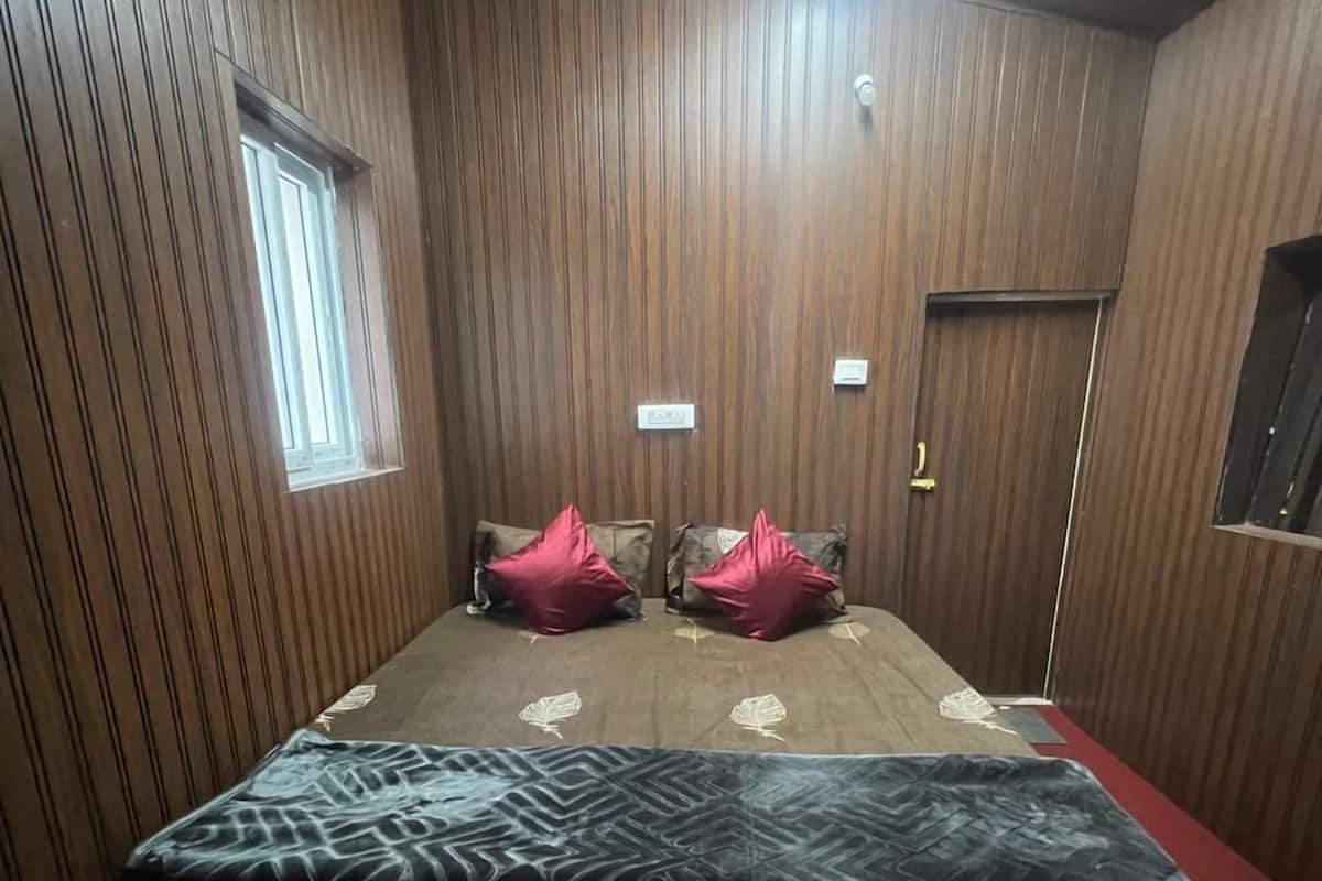 A room with wooden paneling, a bed with a patterned bedspread, two red pillows, a window, and a door.
