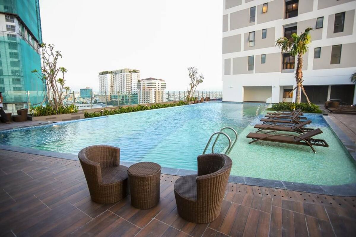 A rooftop pool with wicker chairs and lounge chairs, surrounded by buildings and palm trees.