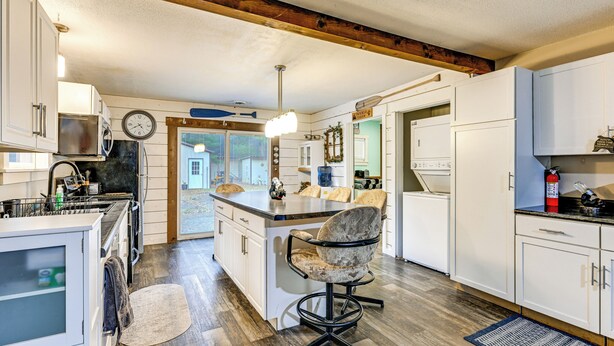 A modern kitchen with white cabinetry, a central island with a chair, a clock on the wall, and a wooden ceiling beam. - 1 Mi to Lake Arbutus: Pet-Friendly Home w/ Games (Merrillan)