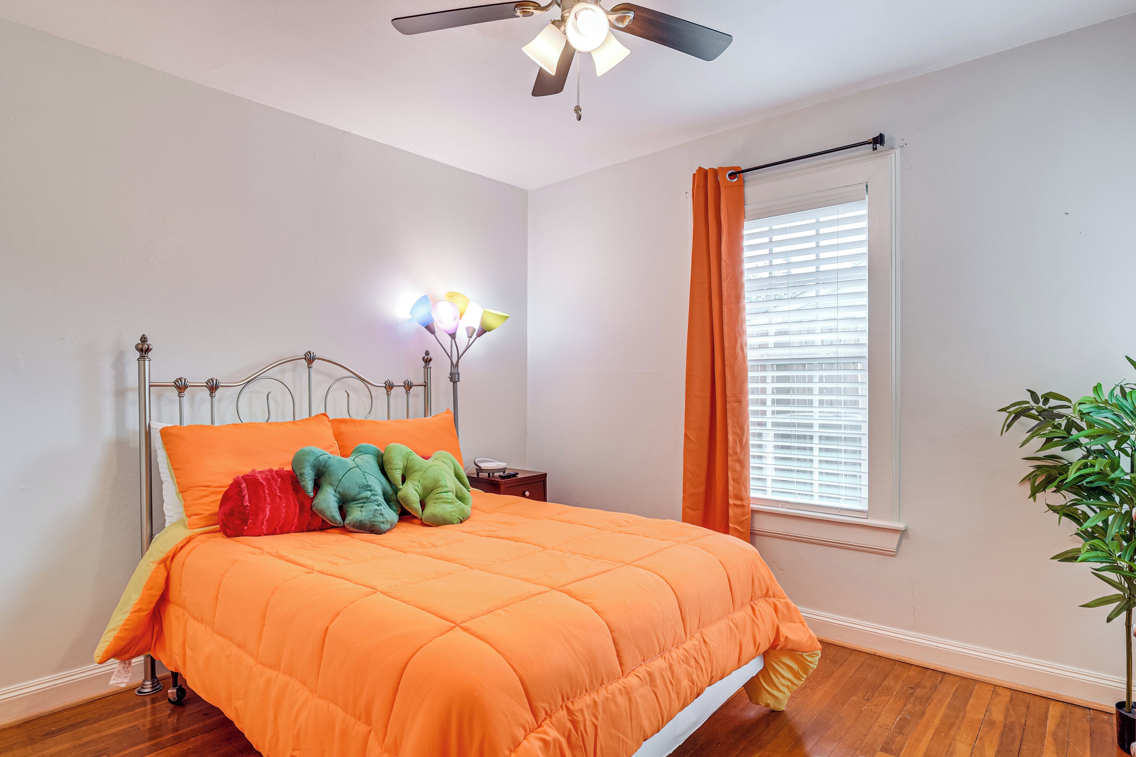 Un dormitorio con estructura de cama metálica, colcha naranja, ventilador de techo, mesita de noche con lámpara y ventana con persianas.