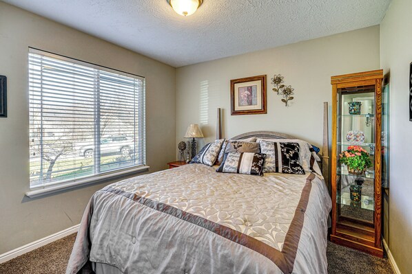 A bedroom with a large bed, a window with blinds, a bedside table with a lamp, and a mirror with a decorative item. - 3,500-Sq-Ft Holiday Gem w/ Game Room in Heber City (Heber City)