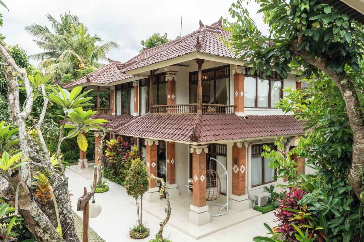 A traditional two-story building with a tiled roof, surrounded by lush greenery and palm trees.