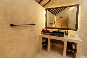 Ein Badezimmer mit Holzbalkendecke, einem großen Spiegel, einem Waschbecken mit rotem Handtuch und Regalen mit Toilettenartikeln.