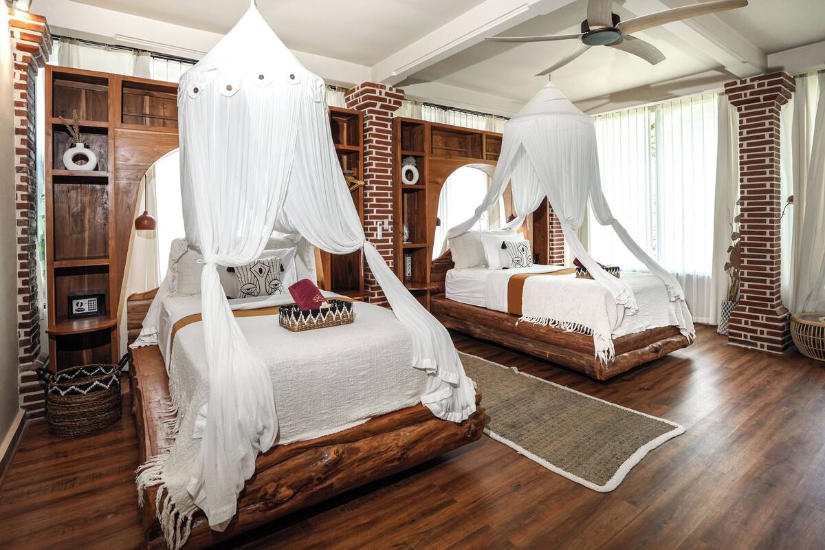A bedroom with two beds, each with a canopy, wooden headboards, and a brick column. There is a wooden wardrobe and a bedside table with a lamp.