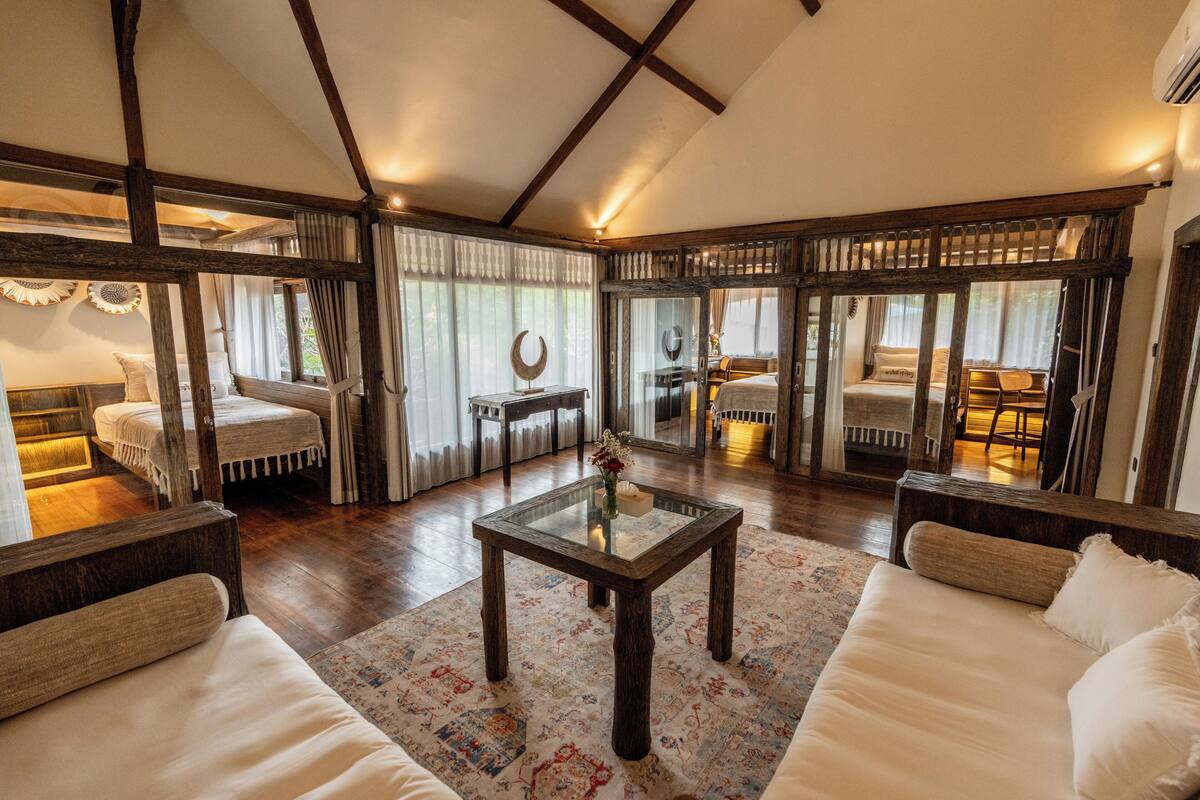 A spacious living room with a high ceiling, wooden beams, a glass coffee table, and a large window with curtains.