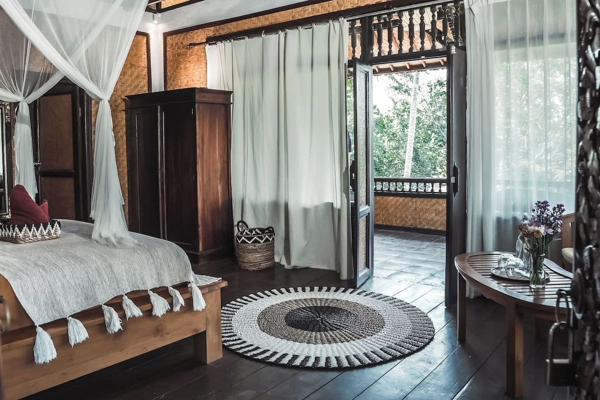 A bedroom with a wooden bed, a wooden wardrobe, a woven basket, a round woven rug, and a balcony with a table and a vase of flowers.