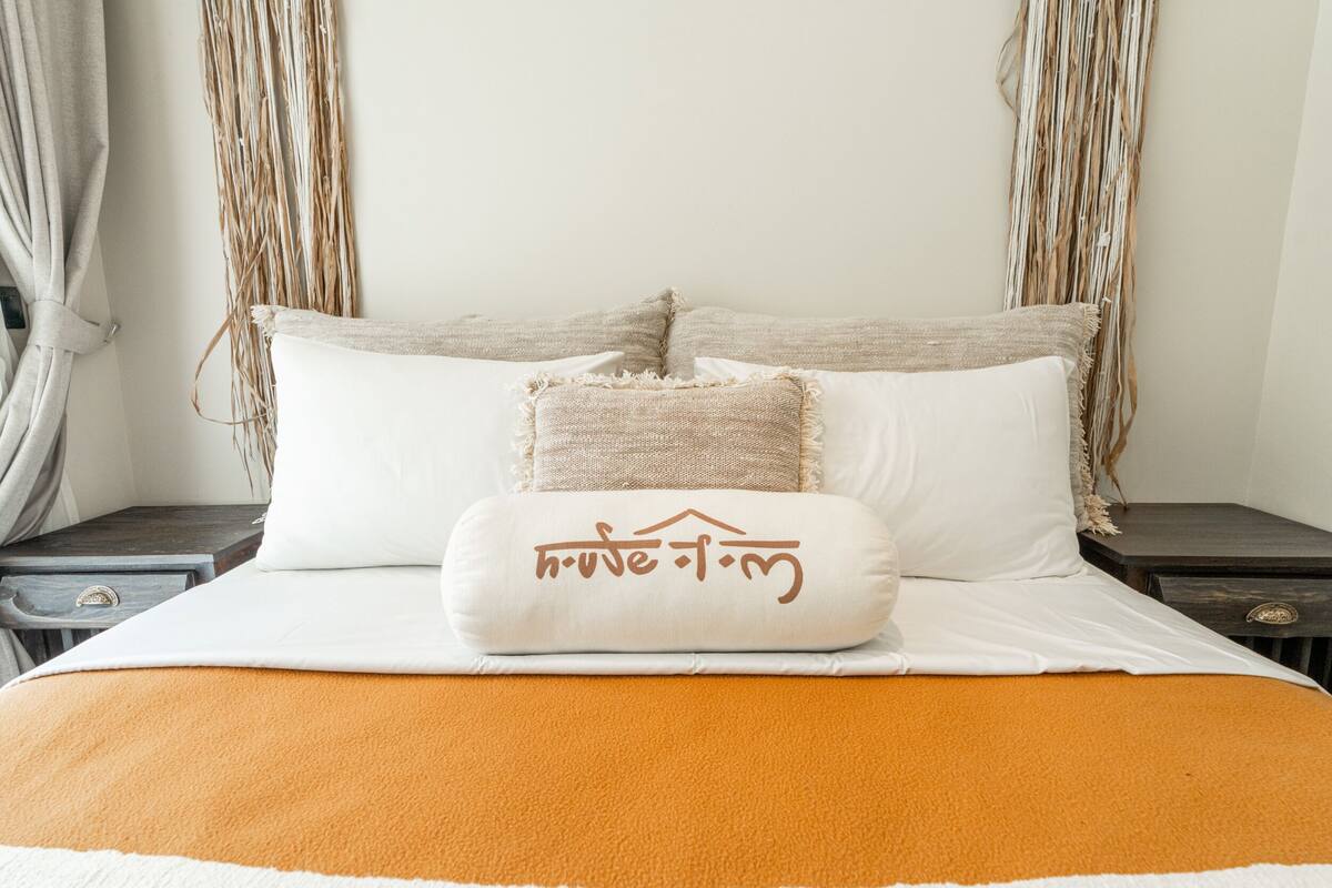 A bed with white and orange bedding, a decorative pillow with text, and a woven wall hanging.