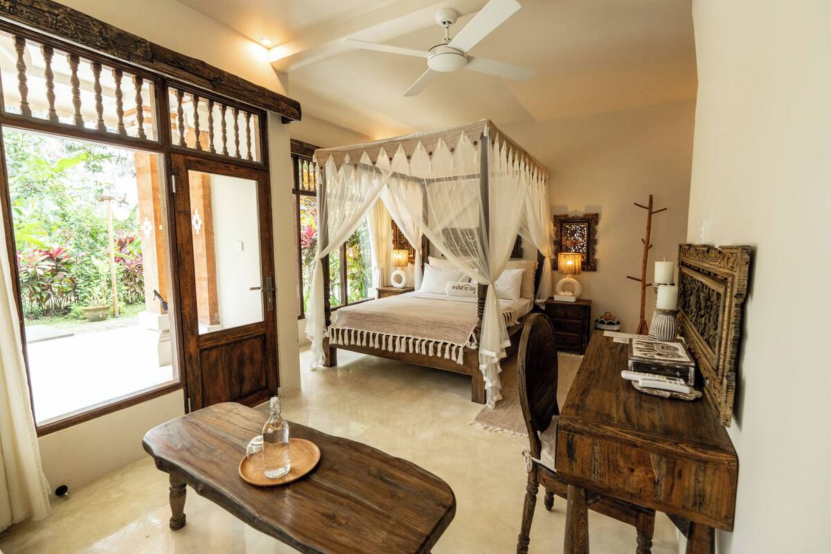 A bedroom with a four-poster bed, a wooden desk, and a view of greenery outside.