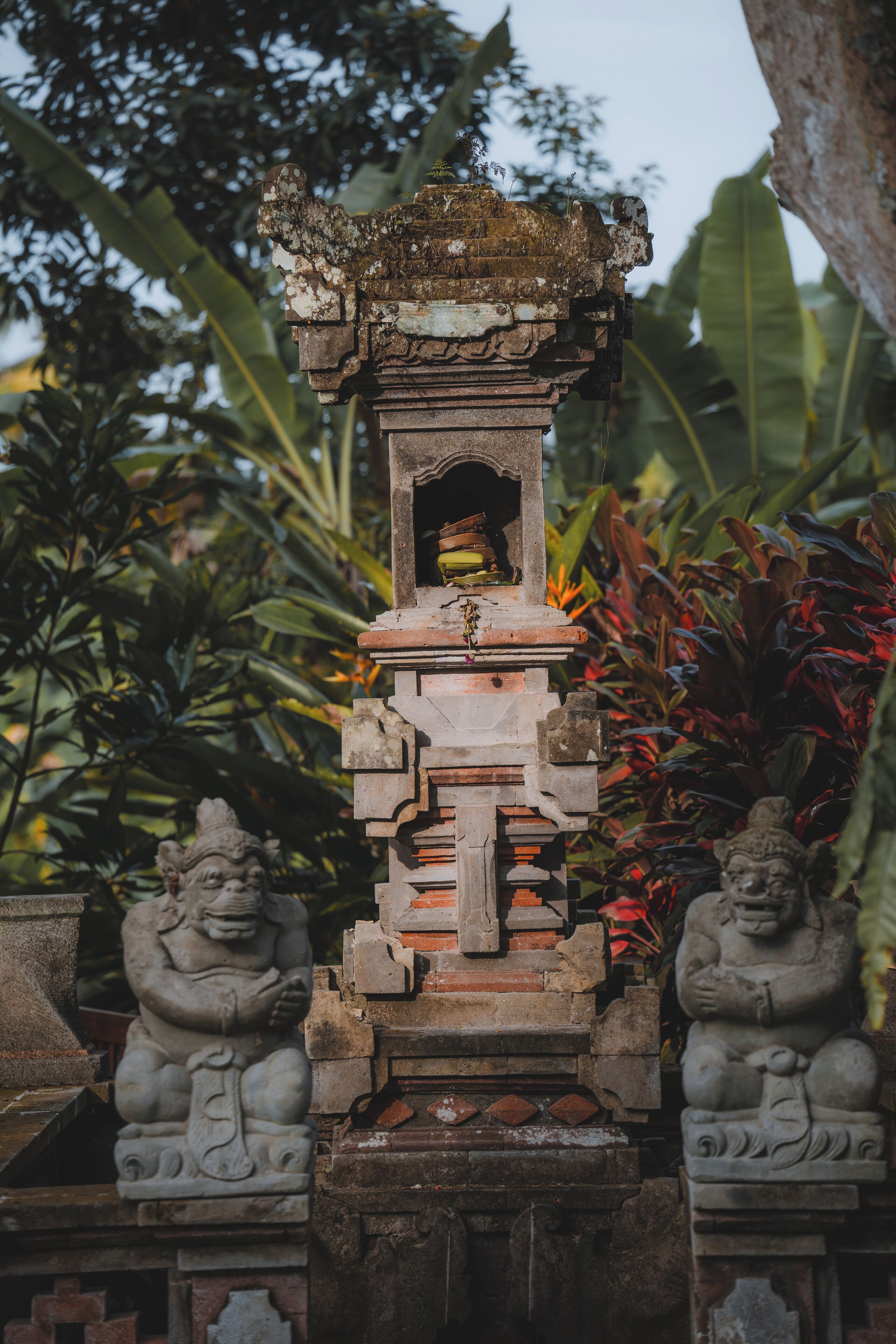 A stone structure with a central doorway, flanked by two stone figures, set against a backdrop of lush greenery.