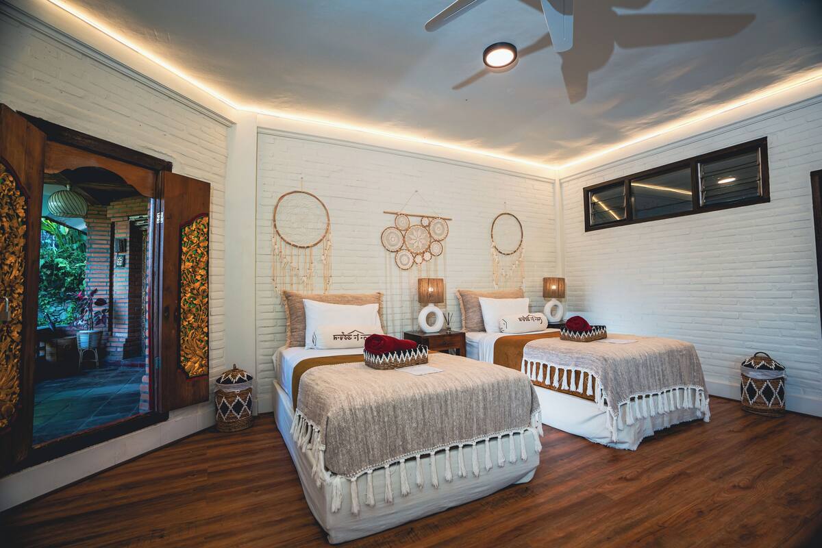 A bedroom with two beds, white brick walls, wooden flooring, and decorative wall hangings.