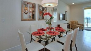 A dining area with a glass table set for six, white chairs, and red napkins. There are abstract paintings on the wall and a large pendant light above the table. - Serenity 3 Bedroom Vacation Home 1523 (Clermont)