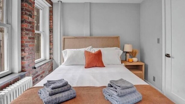 Une chambre d’hôtel avec un mur en briques, un lit avec une literie blanche et une tête de lit en bois, une table de chevet avec une lampe et une fenêtre donnant sur un beau paysage.