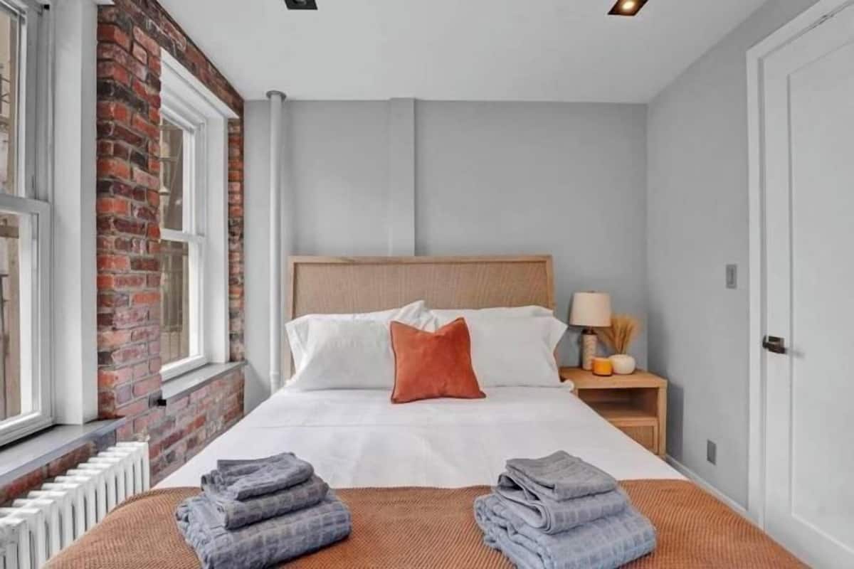 A hotel room with a brick wall, a bed with white bedding and a wooden headboard, a nightstand with a lamp, and a window with a view.