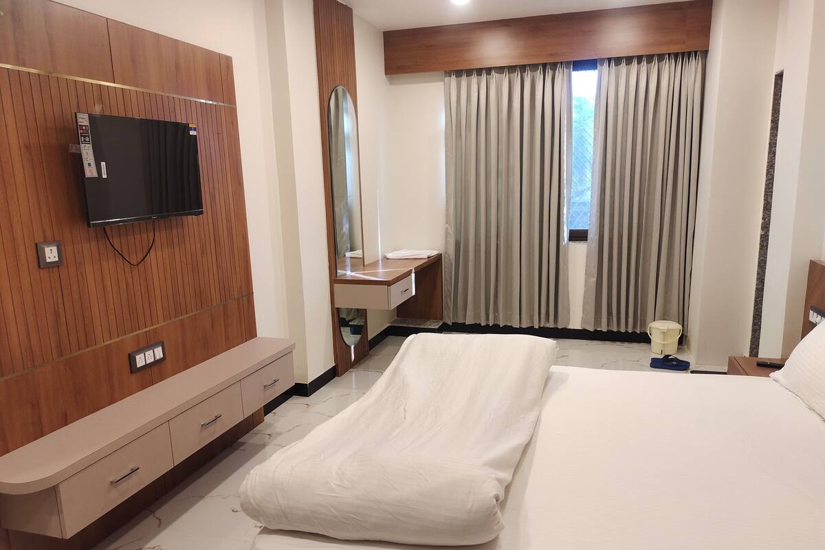 A hotel room with a bed, a TV mounted on the wall, a vanity with a mirror, and a window with curtains.