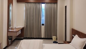 Ein Hotelzimmer mit zwei Betten, einem Deckenventilator, einem Schminktisch mit Spiegel, einem Fenster mit Vorhängen und einem kleinen Tisch mit Eimer.