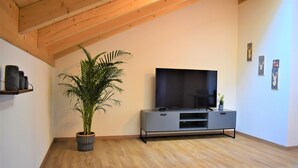 A modern living room with a flat-screen TV on a grey console, a large potted plant, and wooden beams on the ceiling. - Wanderlust apartment with 2 bedrooms, kitchen, shower/WC and loggia (Rosenheim)