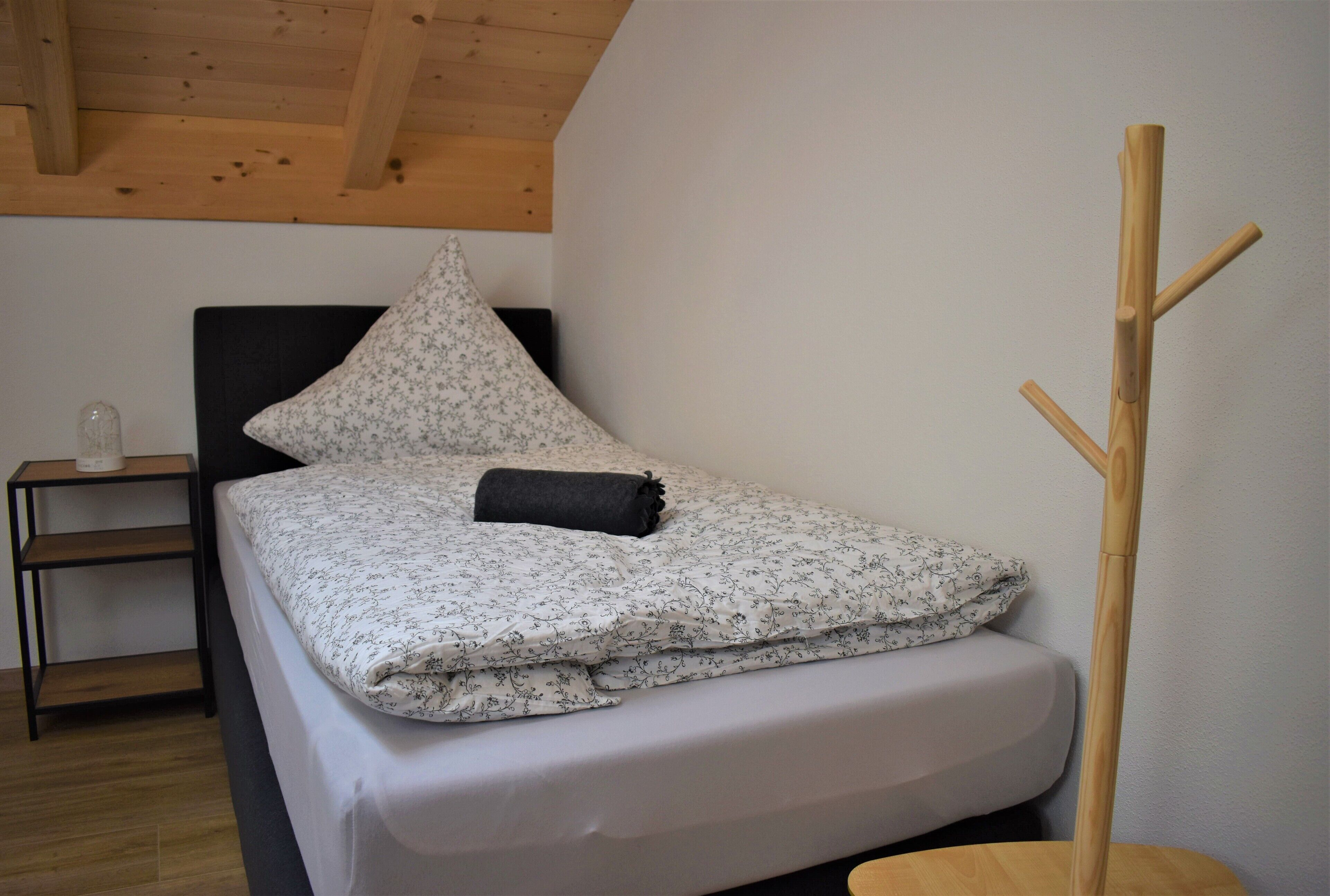 Un lit simple avec une couvre-lit à motifs floraux et un oreiller noir, une table de chevet avec un récipient en verre et un plancher en bois.