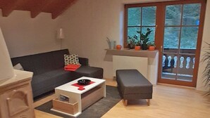 Una sala de estar acogedora con un sofá oscuro, una mesita auxiliar blanca, un reposapiés gris y una ventana con vistas a la vegetación.