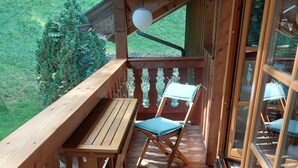 Una terraza de madera con una mesa y dos sillas, un lámpara colgante y vistas a la vegetación.