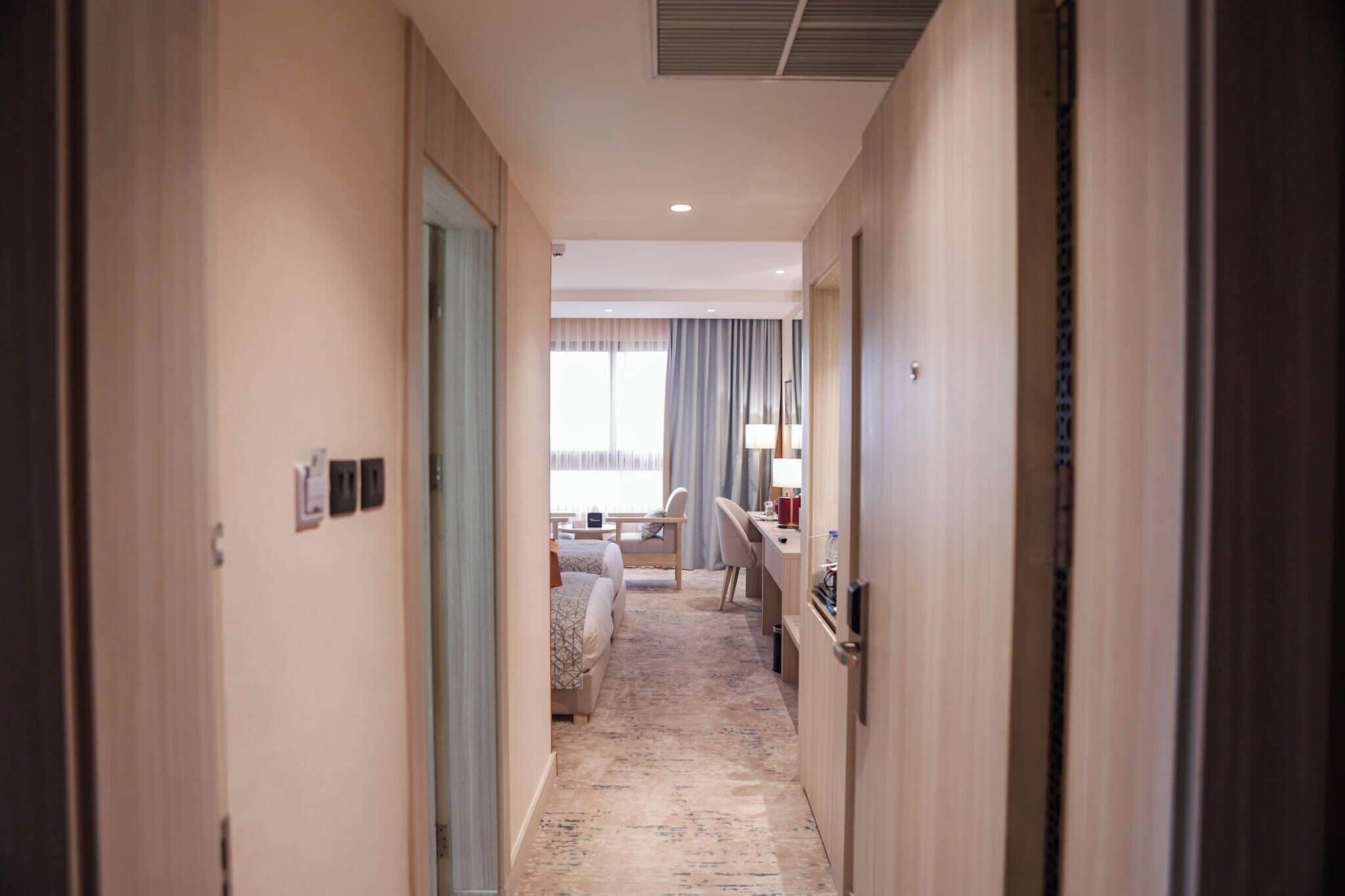 A hotel hallway leading to a bedroom with a bed, a desk, and a chair.
