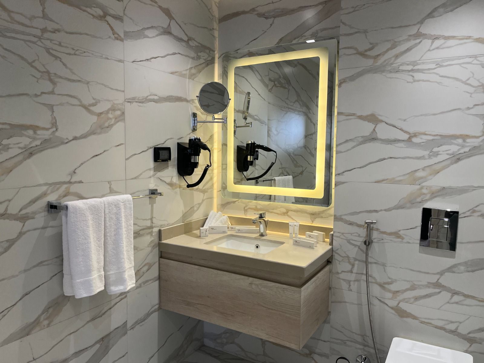 A modern bathroom with marble walls, a wooden vanity, a large mirror, and a towel rack.