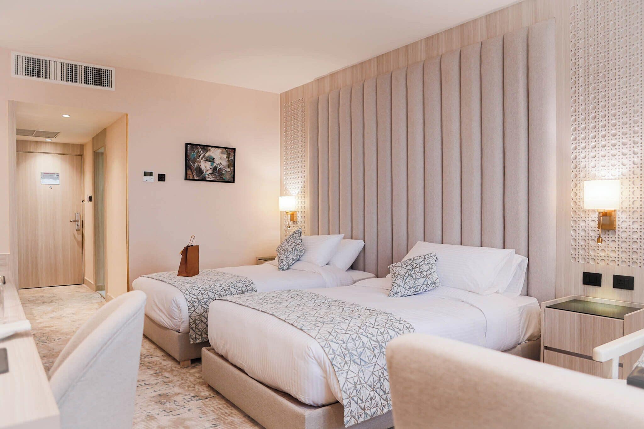 A hotel room with two beds, a desk, a chair, and a wall-mounted lamp.