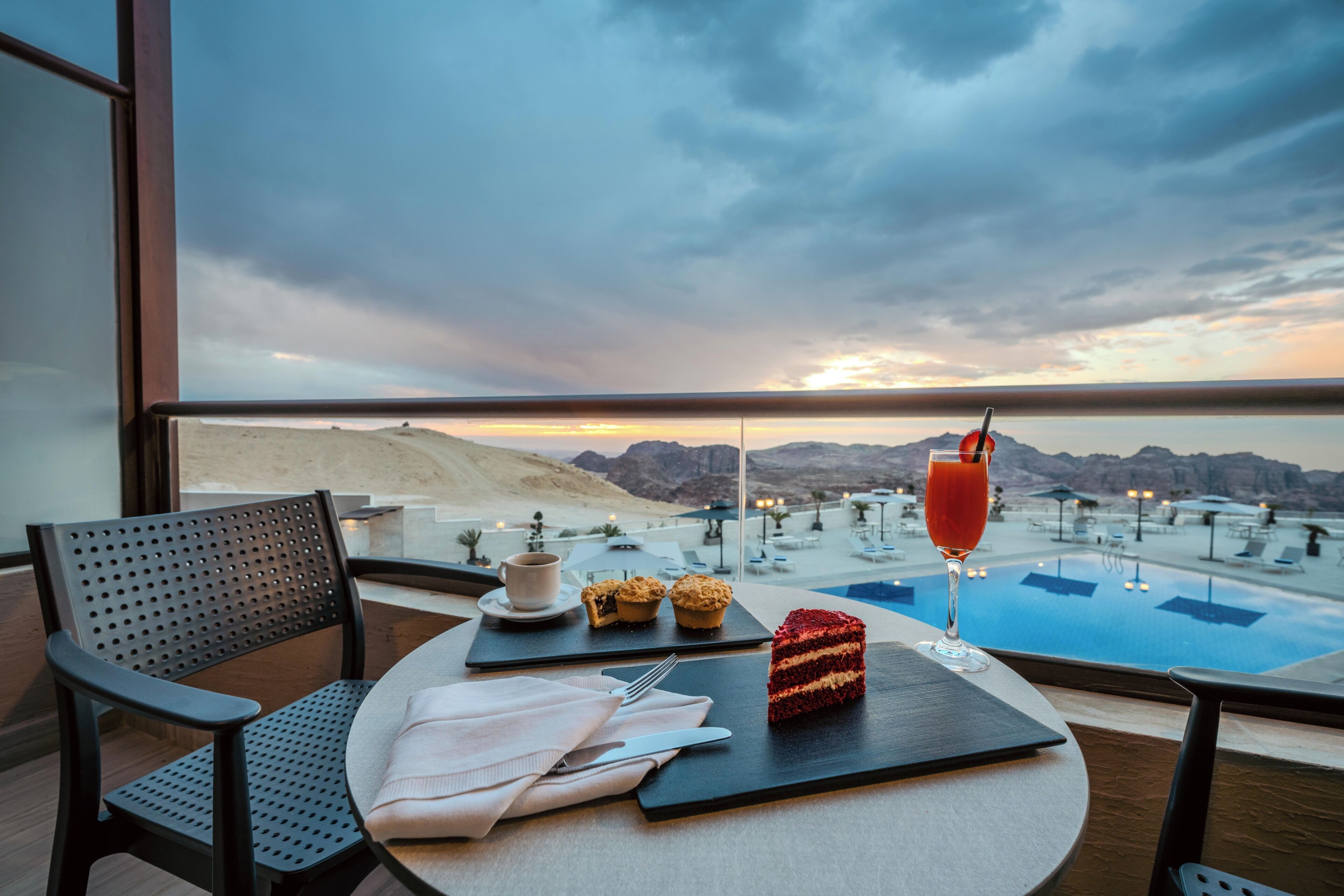Una taula de menjador amb un postre, una beguda i un muffin a un balcó amb vista a una piscina i muntanyes.