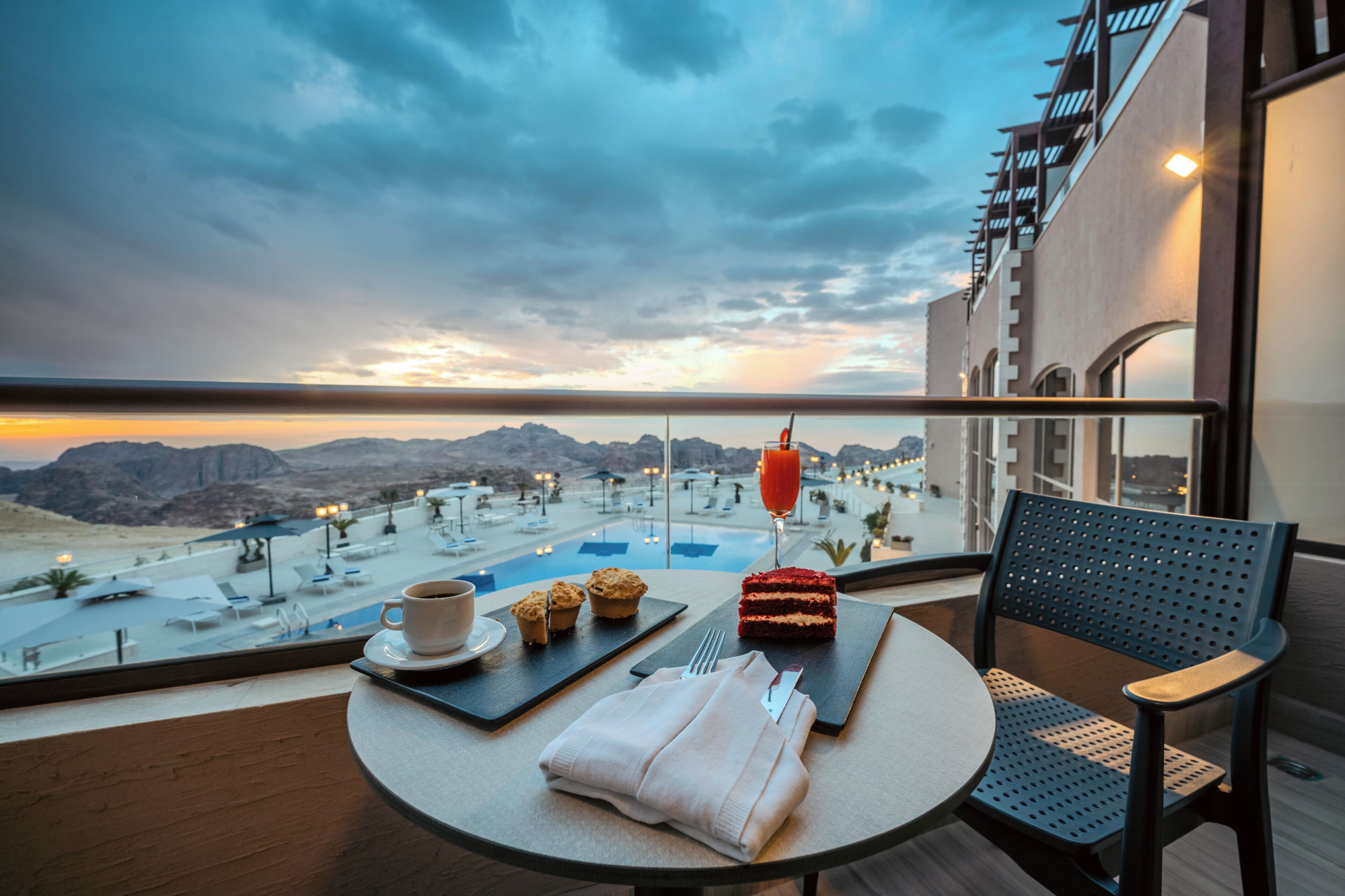 A balcony with a table set for a meal, a chair, and a view of a pool and mountains.