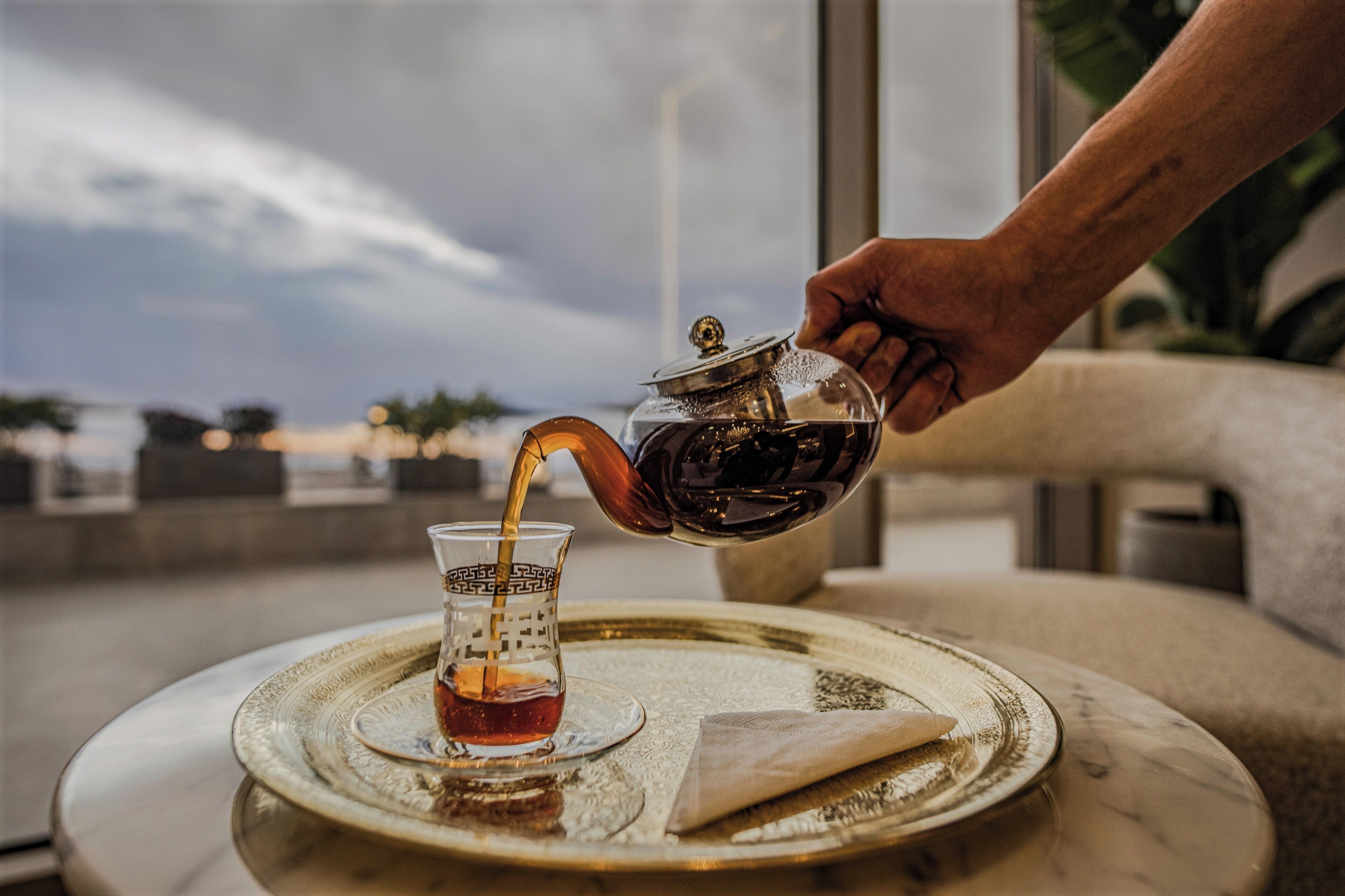 A hand pouring tea into a glass on a tray with a view of the sea in the background.