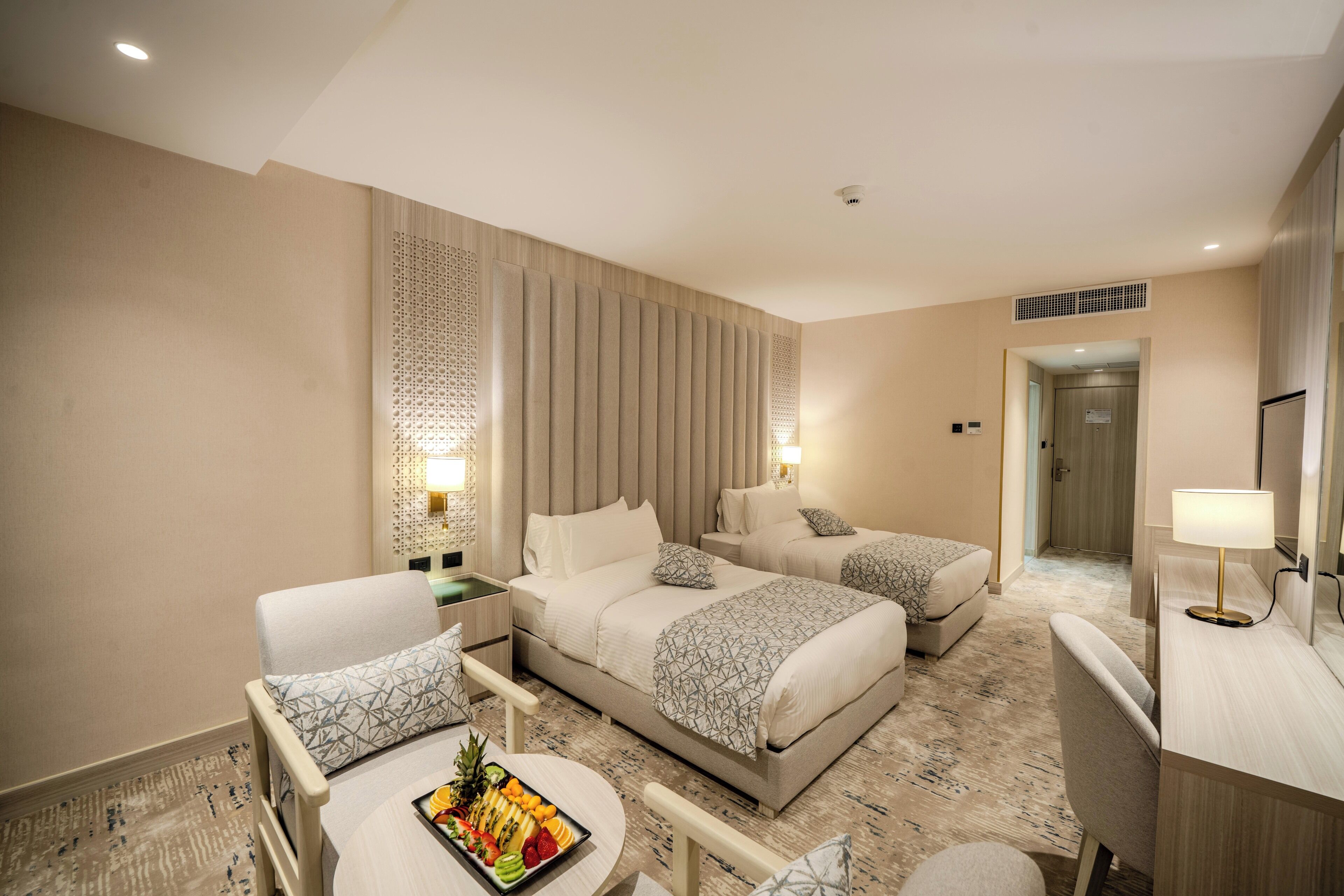 A hotel room with two beds, a desk, a chair, and a small table with fruit.