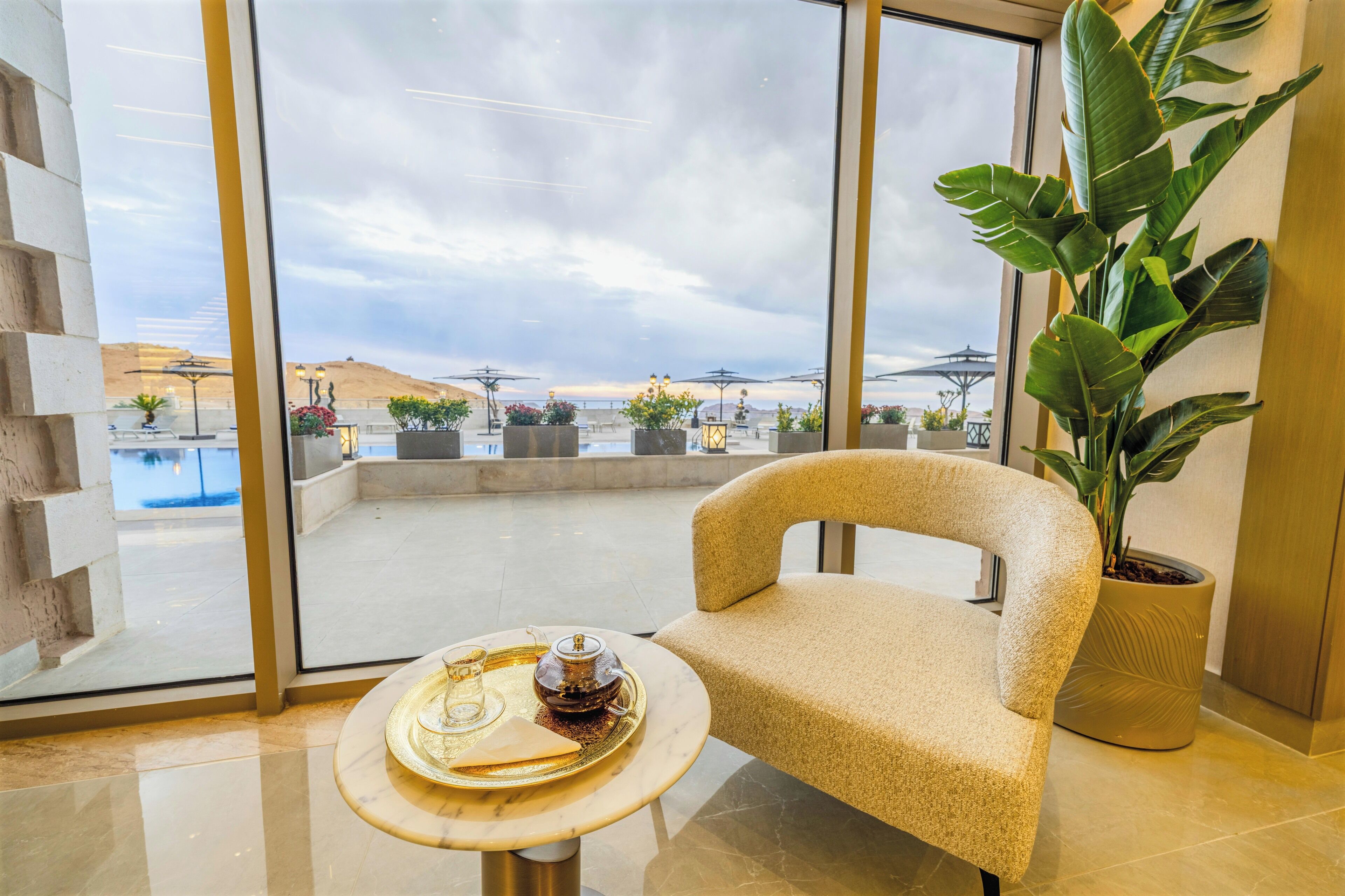 A modern hotel room with a large window, a comfortable chair, a small round table with a tray, and a potted plant.